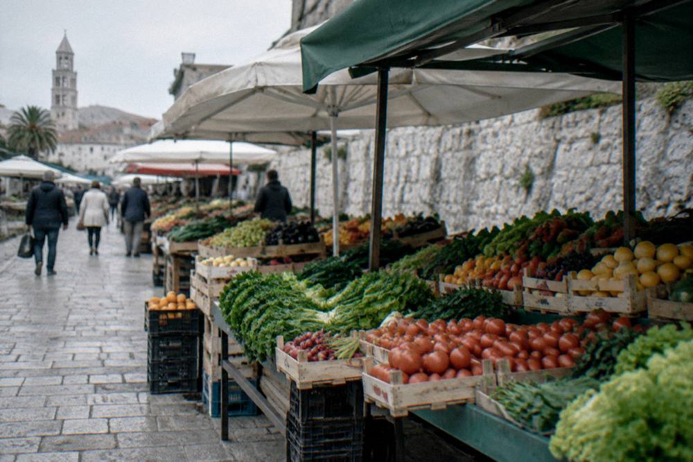 A street market with crates of fruit and vegetables under awnings beside an old stone wall
