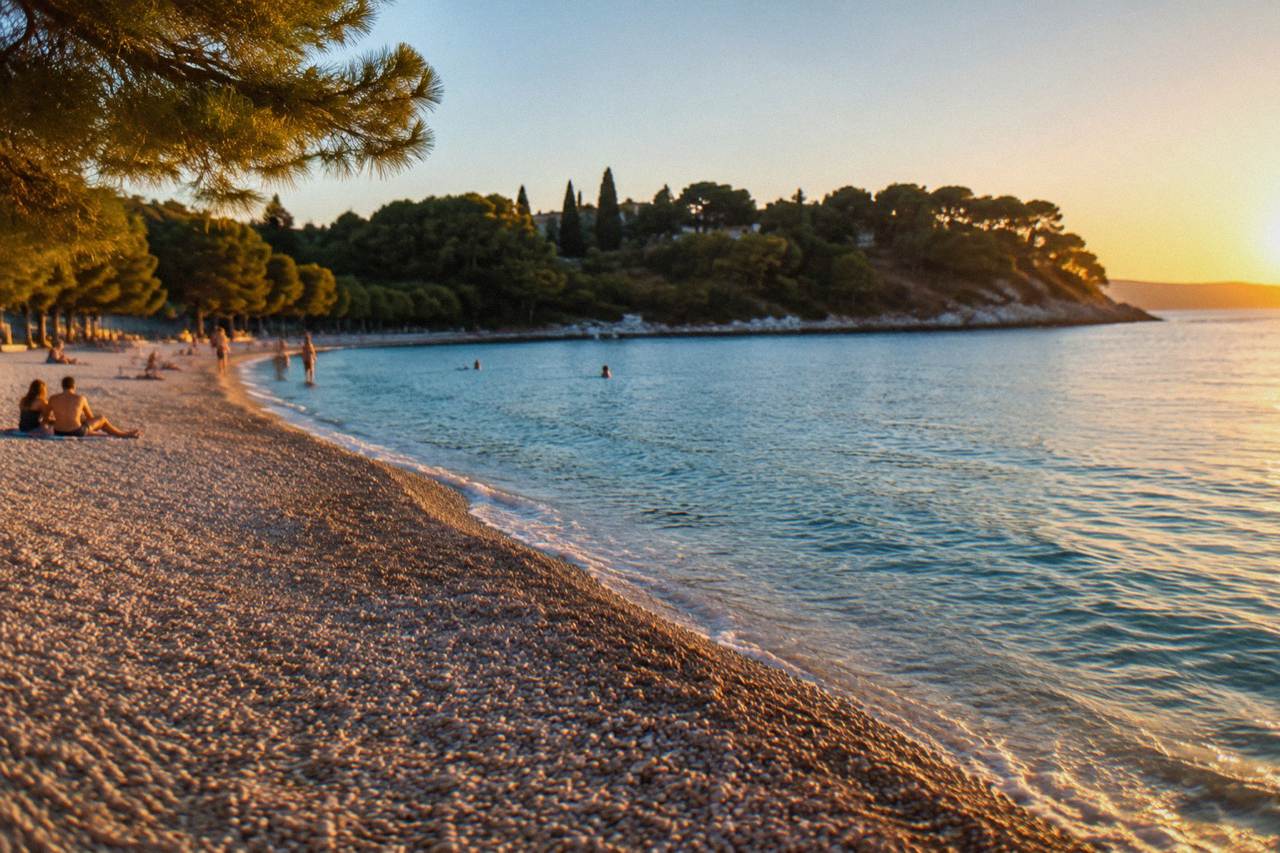 A wide pebble beach curving along a calm blue Adriatic bay with a wooded peninsula behind it