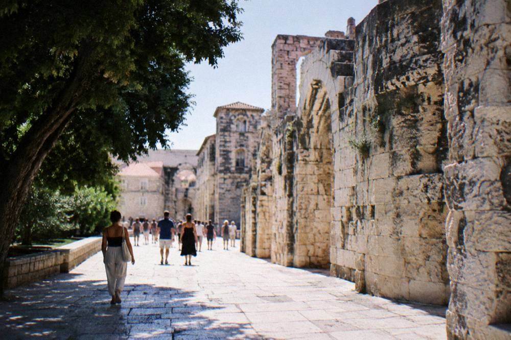 A stone-paved old town street with people in summer clothes walking past a Roman wall