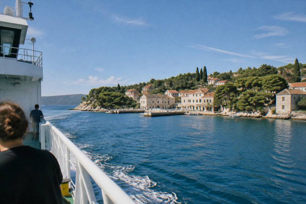 Car ferry approaching an island port town with pine trees and stone houses along the shore