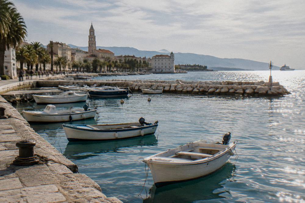 Sun glinting on the sea beyond a stone breakwater with rowboats moored in a small Dalmatian harbour