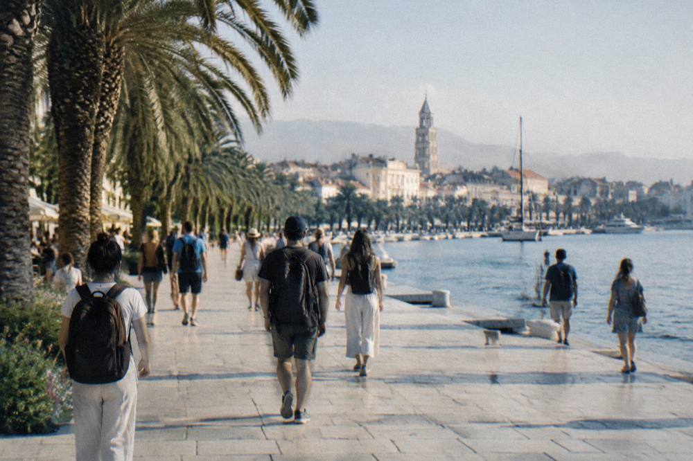 Crowd of travellers with day bags walking along a sunny waterfront promenade lined with palm trees
