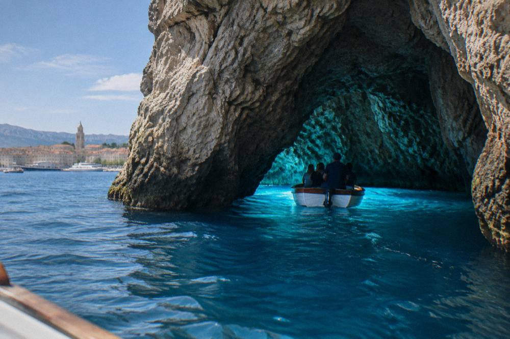 Small wooden boat entering a glowing blue sea cave lit from below the waterline
