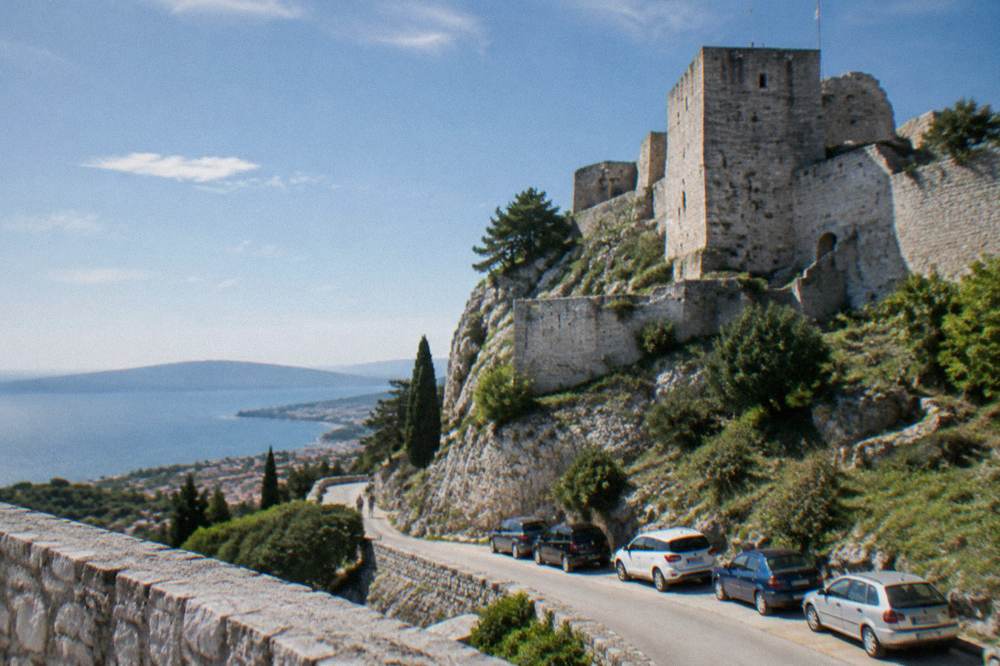A stone fortress on a hill above a coastal road with parked cars below