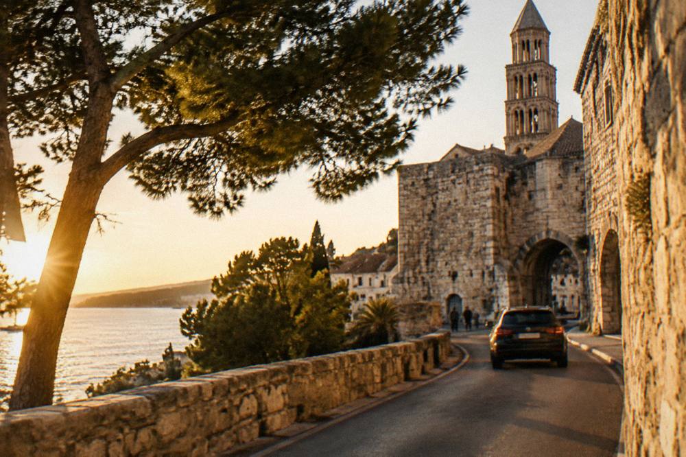 A car on a sunny coastal road with dry stone walls and pine trees