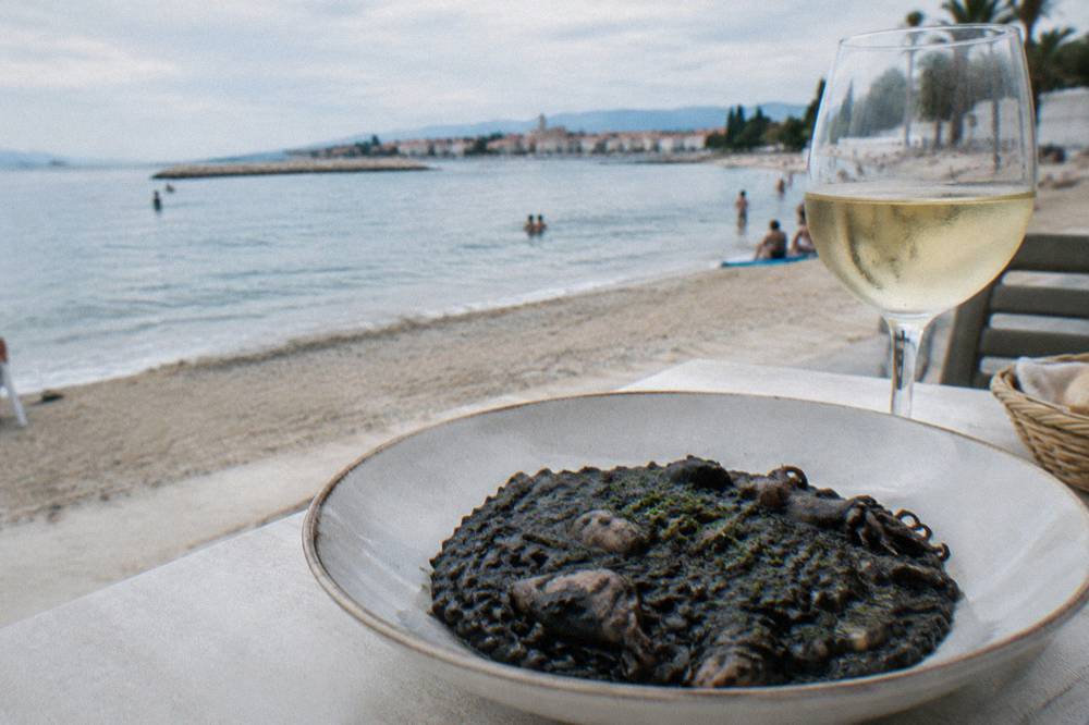 A dark cuttlefish ink risotto served in a shallow bowl beside a glass of white wine