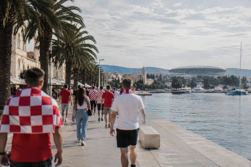 Fans in red and white walking along a seaside promenade toward a stadium