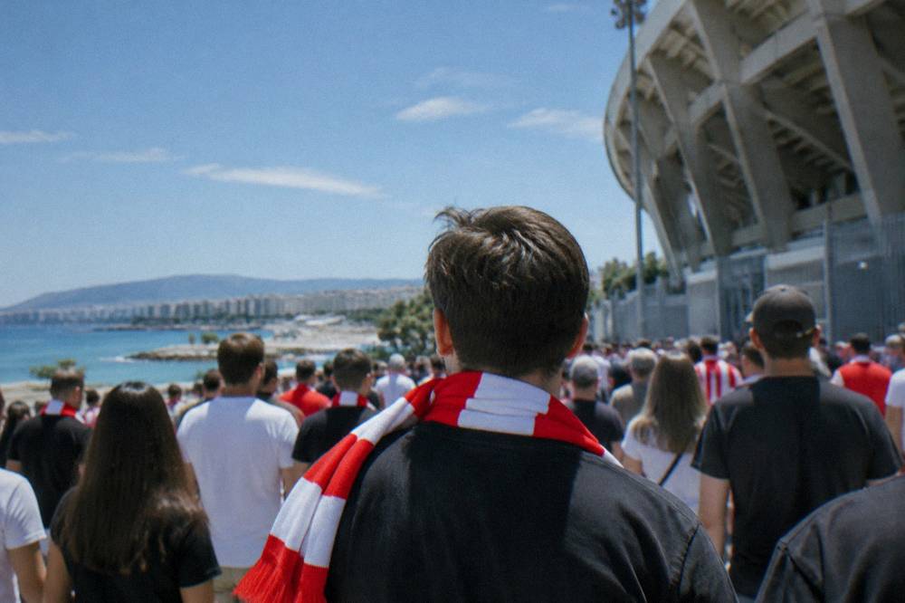 A supporter with a red and white scarf standing among a crowd outside a stadium gate