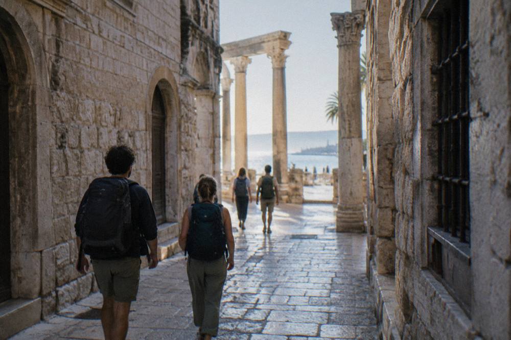 Travellers with backpacks walking a narrow stone street in Split toward sunlit pillars
