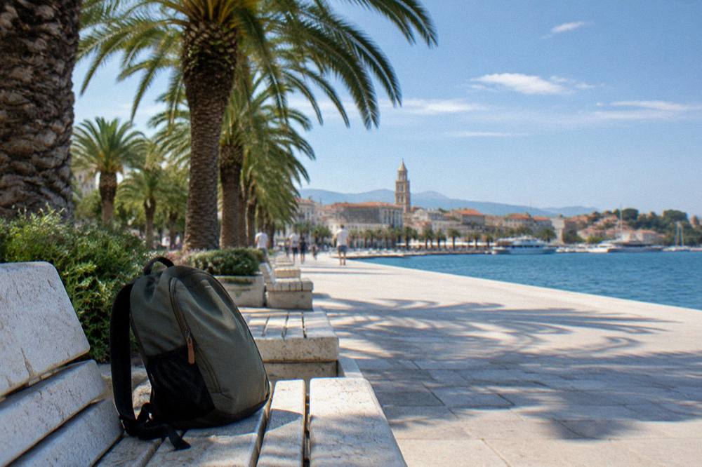 A small daypack on a stone bench beside the seafront promenade in Split with palm trees above