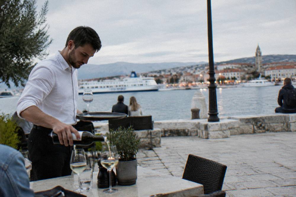 Waiter serving wine on a stone terrace with harbour lights beyond