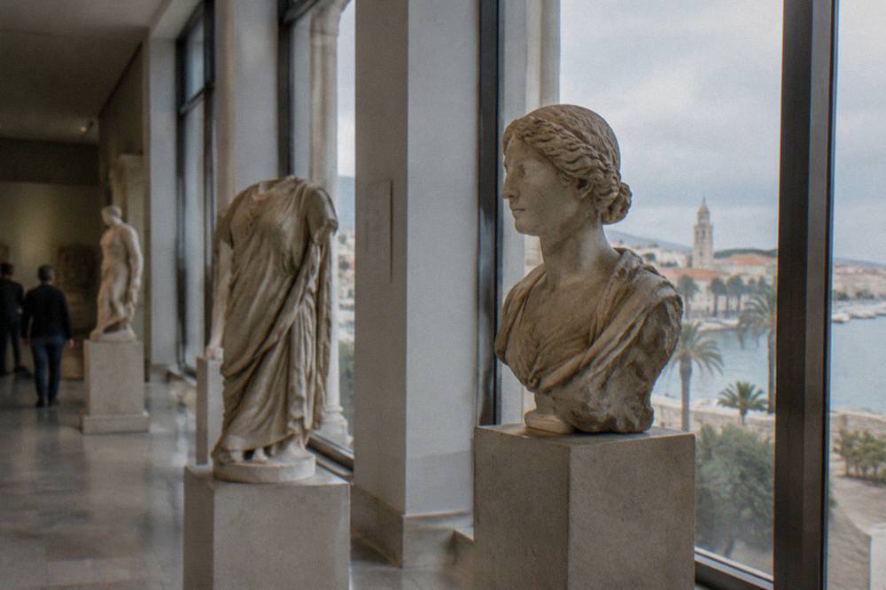 A gallery interior with sculptures on plinths beside tall windows