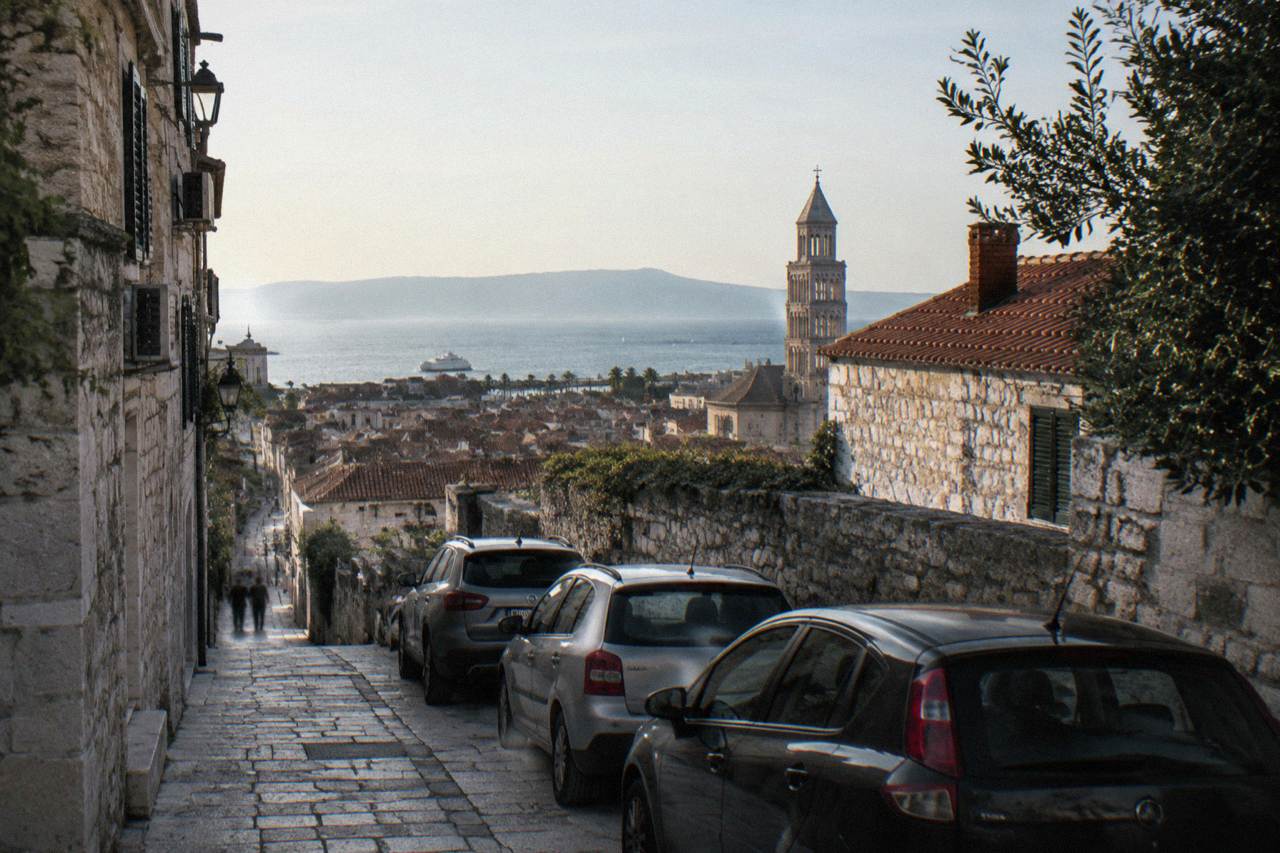 Cars parked in a narrow stone street above the rooftops of Split with the Adriatic sea in the distance