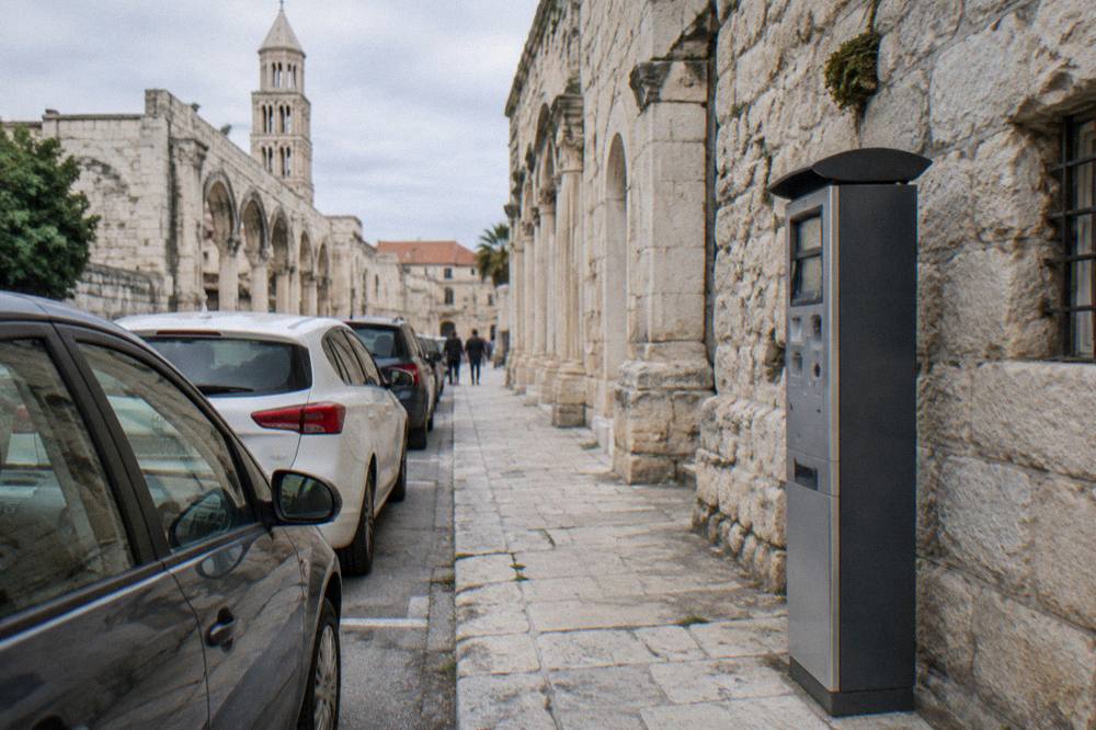 A pay-and-display ticket machine on a sunny street in Split with parked cars in a row