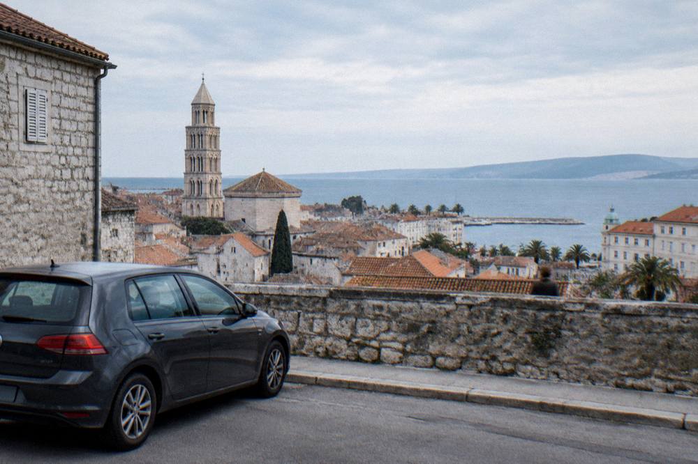 A parked car on a coastal road with a view over stone rooftops and the blue sea near Split