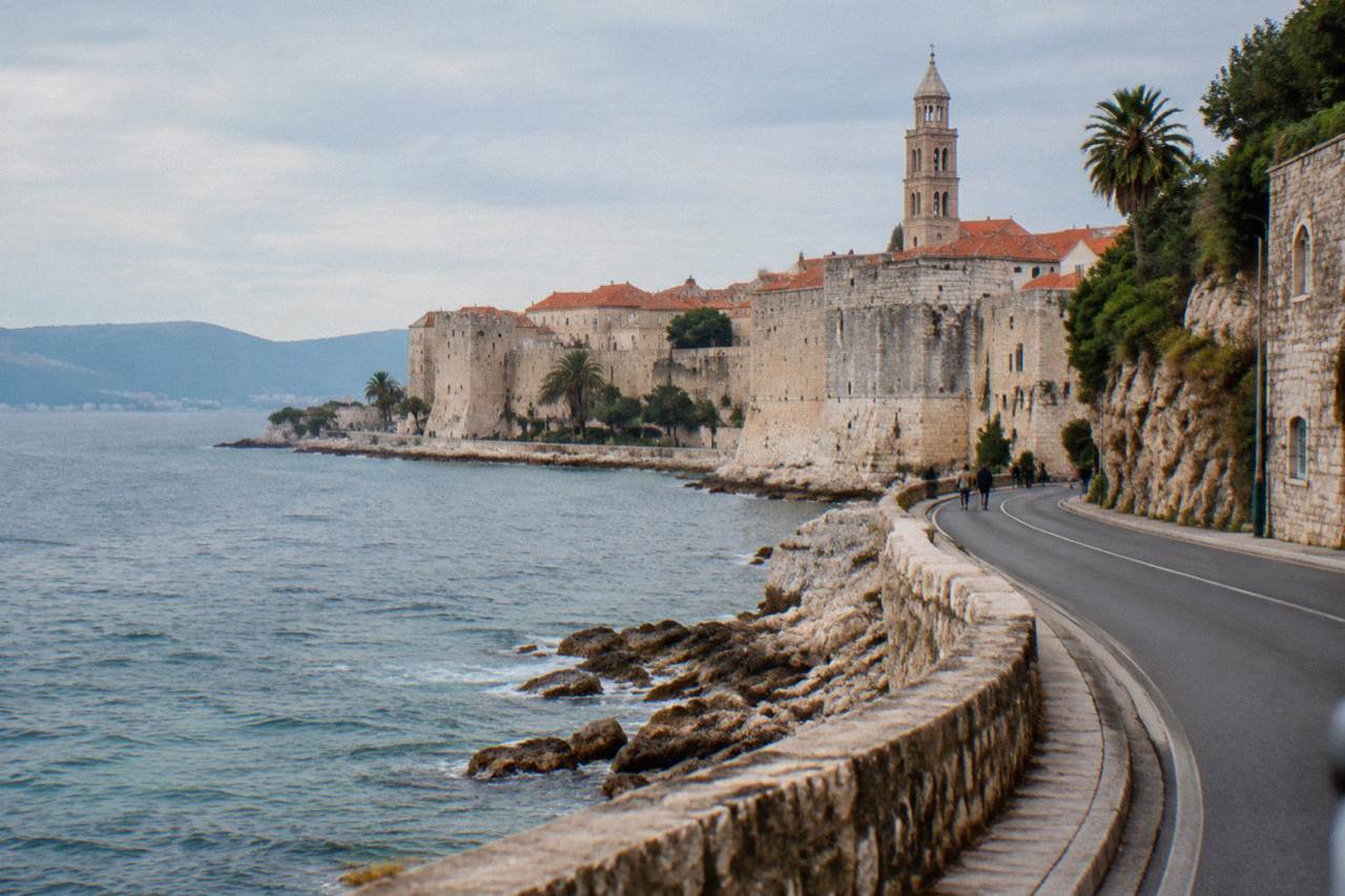 A coastal road winding along the Adriatic Sea toward a walled old town on a rocky shoreline