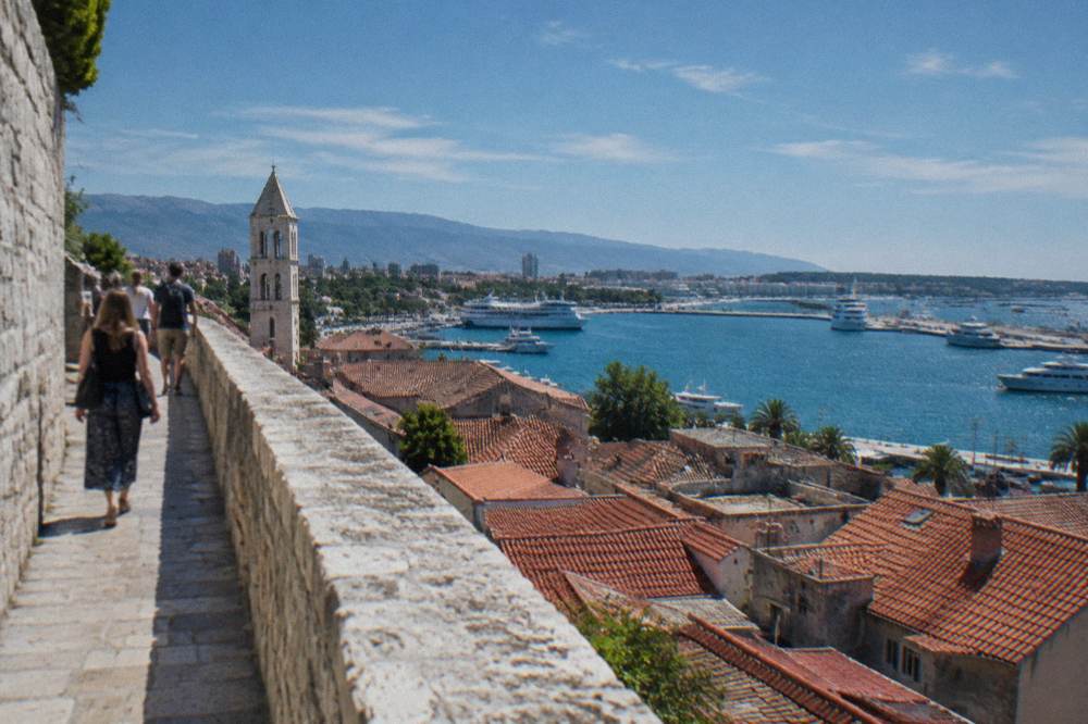 Visitors walking a high stone wall path above red rooftops and a blue harbour