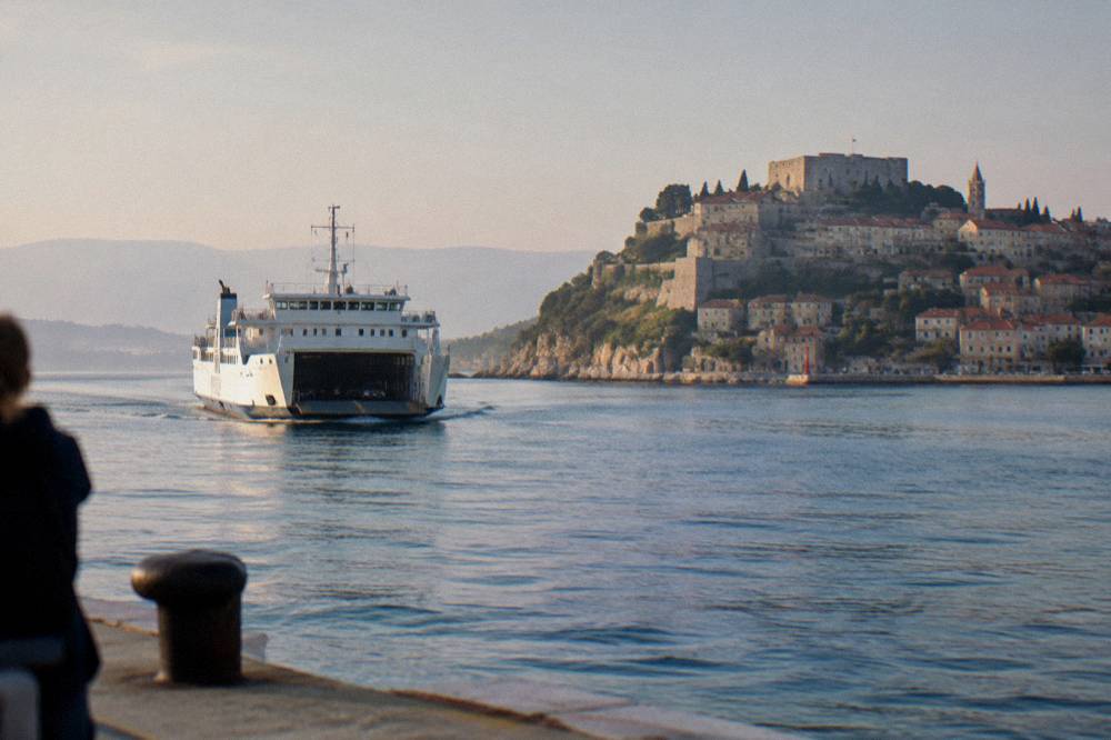 A car ferry approaching an island harbour with a fortified town on the hillside behind