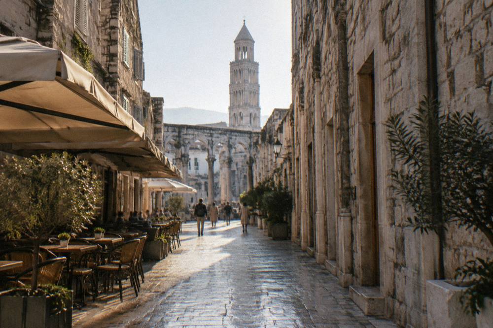 A sunlit stone street between tall old buildings with cafés and a few pedestrians