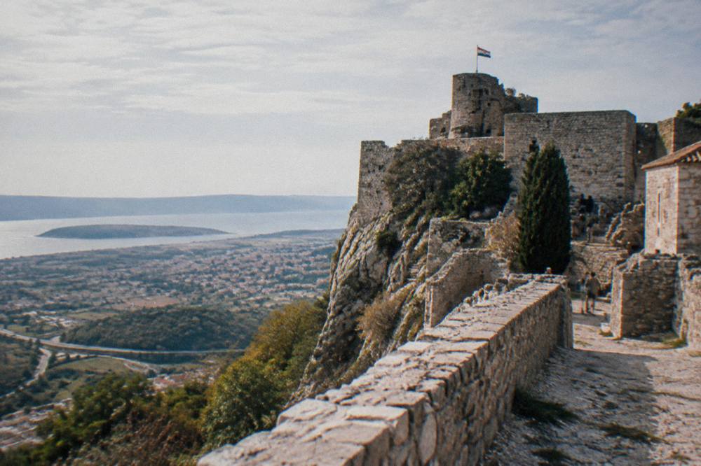 A medieval fortress on a cliff ridge overlooking a coastal plain and distant sea