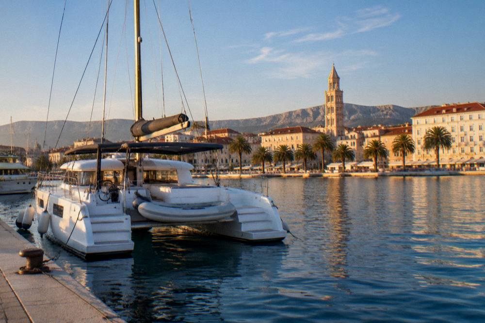 A catamaran moored in a marina with a historic waterfront town behind it at golden hour
