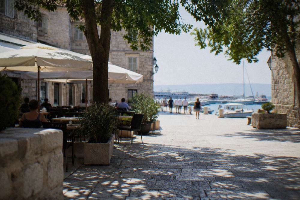 A stone-walled town square with cafes and a harbour with small boats beyond