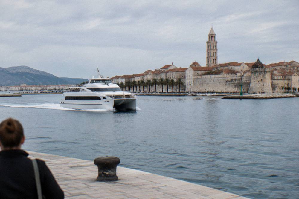 A white catamaran speeding past a walled Adriatic old town on a clear morning