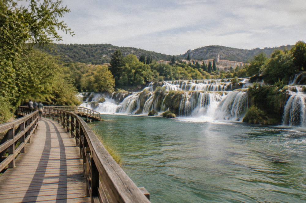 Wide wooden boardwalk crossing green water above a broad travertine waterfall