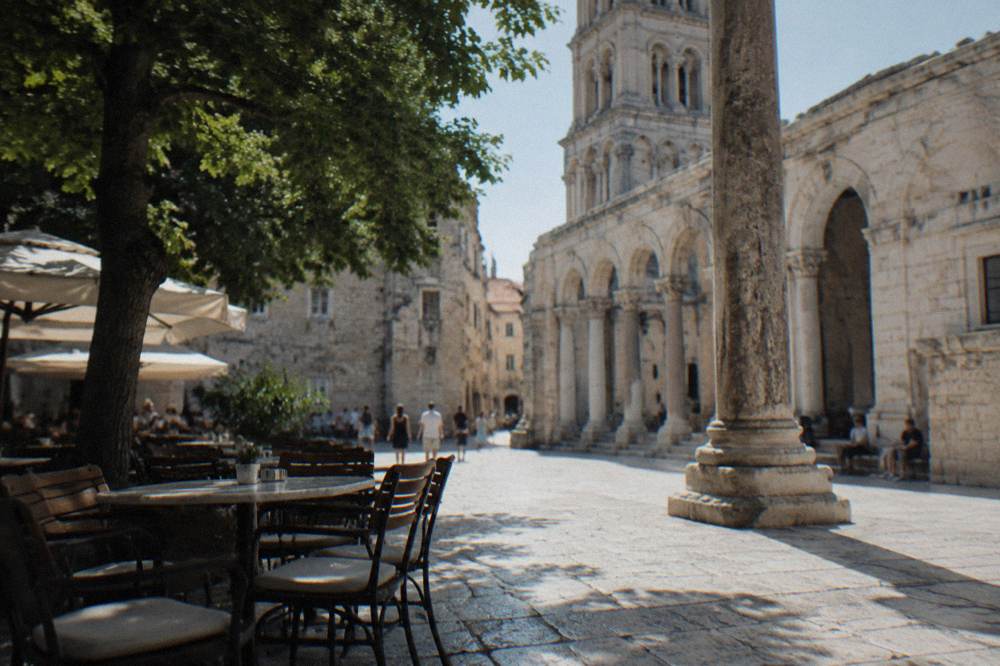 Stone paved square by a Roman column with cafe tables under warm afternoon light