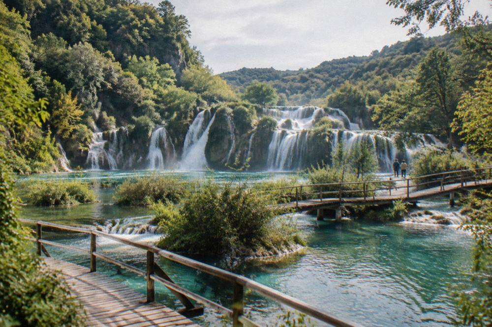 Wooden boardwalks crossing turquoise lakes beneath waterfalls in a forested national park