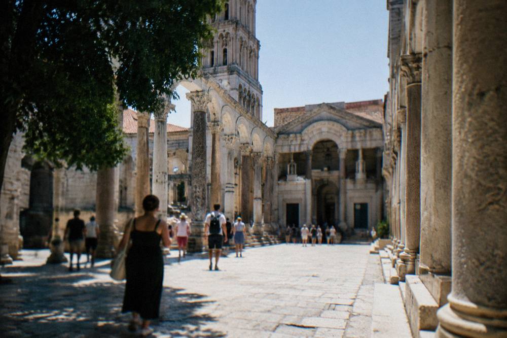 A stone palace courtyard with roman columns and people walking through