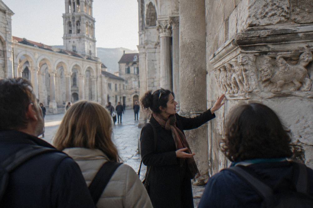 A guide explaining stone carvings to a small group of visitors on a walking tour in an old town square