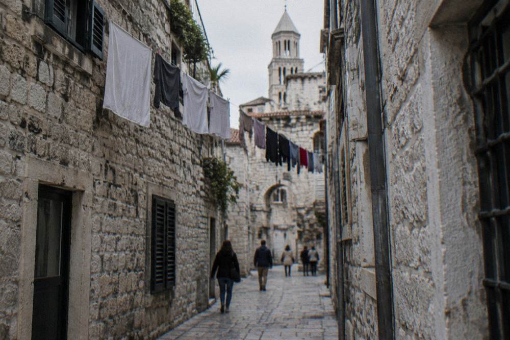 People walking through a narrow stone street in the old town with laundry overhead
