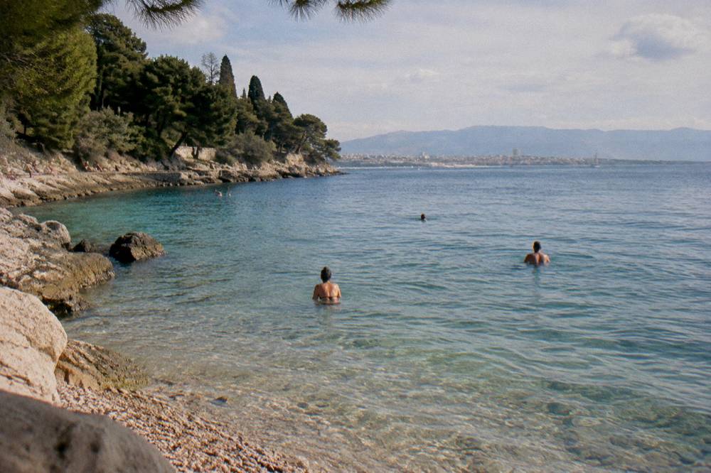 Swimmers in the clear shallow water at a pebble beach below Marjan