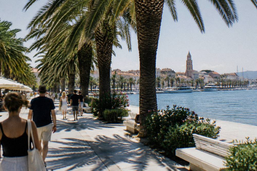 People strolling on the wide Riva waterfront promenade in Split beside palm trees